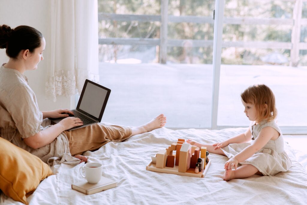 Why multitasking doesn't work - mom sitting on bed and surfing laptop with blank screen while adorable kid playing with toys in cozy bedroom